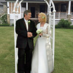 Colour photo of man and woman outside under an arch wearing wedding dress and suit