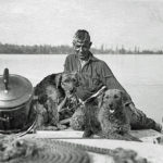 B&W photo of man with two dogs on a boat on lake